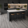 A modern kitchen island with a white stone countertop and grey base cabinet, styled with three wooden-topped bar stools. The kitchen features dark gray cabinets, a light stone backsplash, and gray wood-look floor tiles.