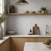 A modern kitchen interior with light grey square mosaic backsplash, white marble countertops, light wood cabinetry, a wooden floating shelf with ceramic dishes and plants, and a gold faucet.