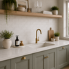 A modern kitchen scene with a matte brass gooseneck faucet installed above a white double-basin sink, set into sage green cabinets with brass knobs. A white subway tile backsplash is visible behind the sink, with a light wood floating shelf and decorative plants on the shelf and countertop.