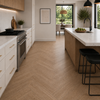 A modern kitchen interior with light oak herringbone wood flooring, white cabinets, a stainless steel gas range, a marble-topped island, and two black bar stools.