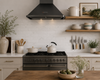 A modern kitchen scene featuring a black range hood mounted above a black gas stove, with white marble subway tile backsplash, light wood floating shelves, cream cabinetry, and kitchenware including a white kettle and ceramic pots.
