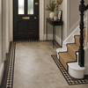 A home entryway featuring large light beige stone-look floor tiles with a black and white geometric border. To the right, a staircase with beige carpet and white steps is bordered by the same tile pattern.