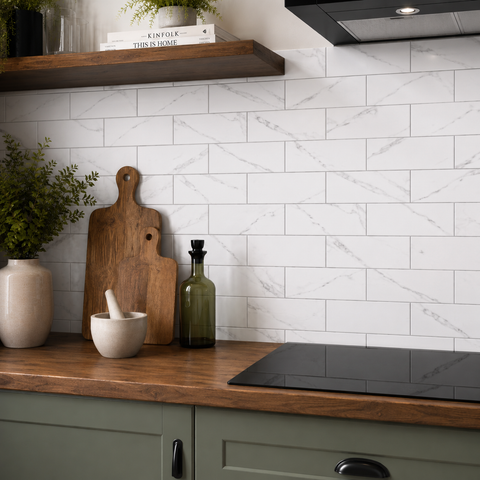 White subway tile backsplash with gray marble veining, installed on a kitchen wall above a wooden countertop with sage green cabinets, a black induction cooktop, a wooden cutting board, and decorative bottles.