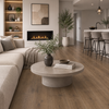 A modern living room with light oak wood plank flooring, featuring a beige sectional sofa, a round concrete coffee table, a black fireplace, and a kitchen area in the background.